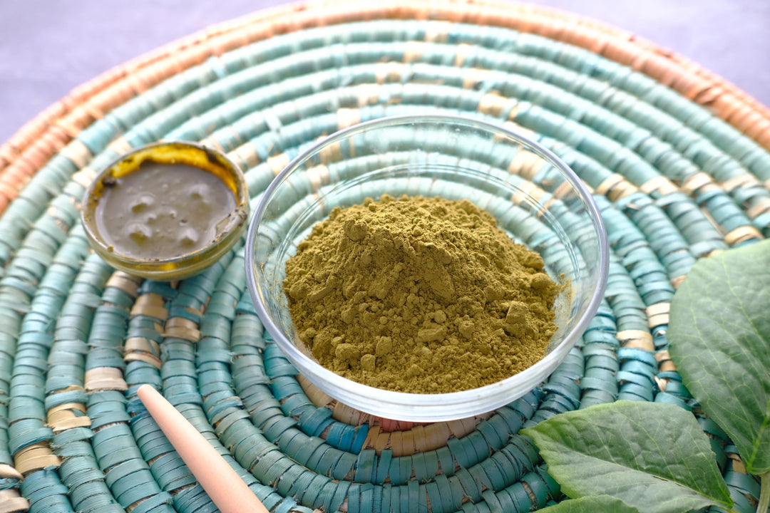 oudre de henné naturelle dans un bol en verre, accompagnée d'une feuille de henné et d'un récipient contenant du henné mélangé, le tout posé sur un panier tissé aux couleurs pastel.