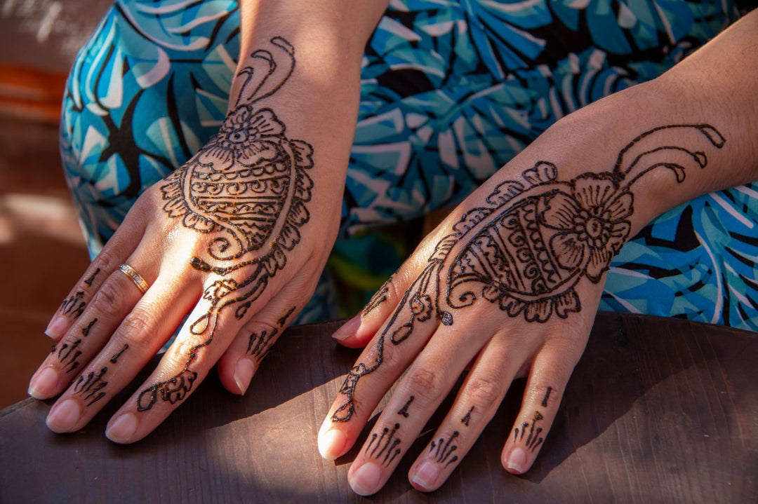 Deux mains présentant des motifs de henné traditionnels avec des détails paisley et floraux, posées sur un fond aux motifs géométriques bleus.