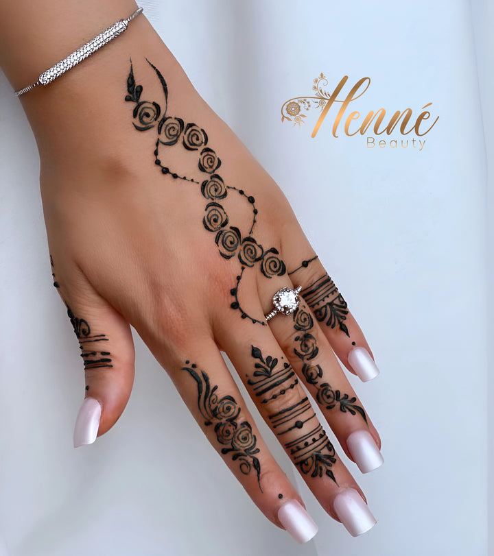 Main féminine affichant un tatouage au henné noir avec des motifs en spirale et linéaires, complété par des ongles vernis en rose pâle et orné d'un bracelet en argent et d'une bague en diamant scintillant