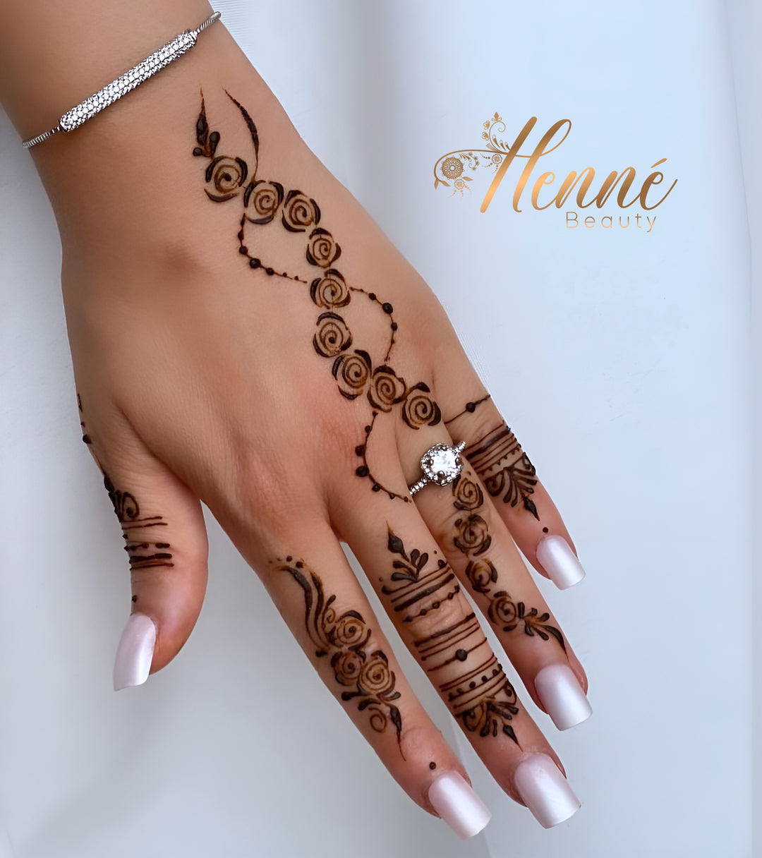 Main féminine affichant un tatouage au henné noir avec des motifs en spirales et linéaires, accentué par des ongles vernis de couleur rose pâle et un bracelet en diamant étincelant.